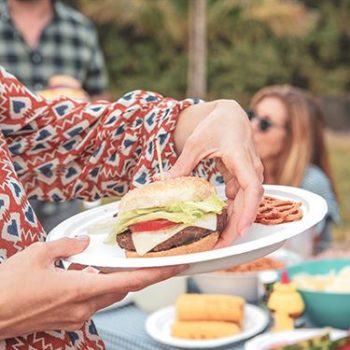 A woman at an outdoor barbecue picking a cheeseburger up off a Chinet Classic plate