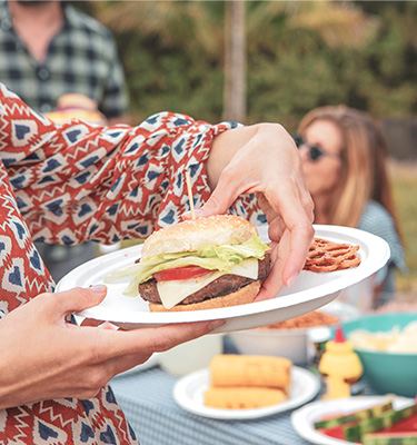 A woman at an outdoor barbecue picking a cheeseburger up off a Chinet Classic plate