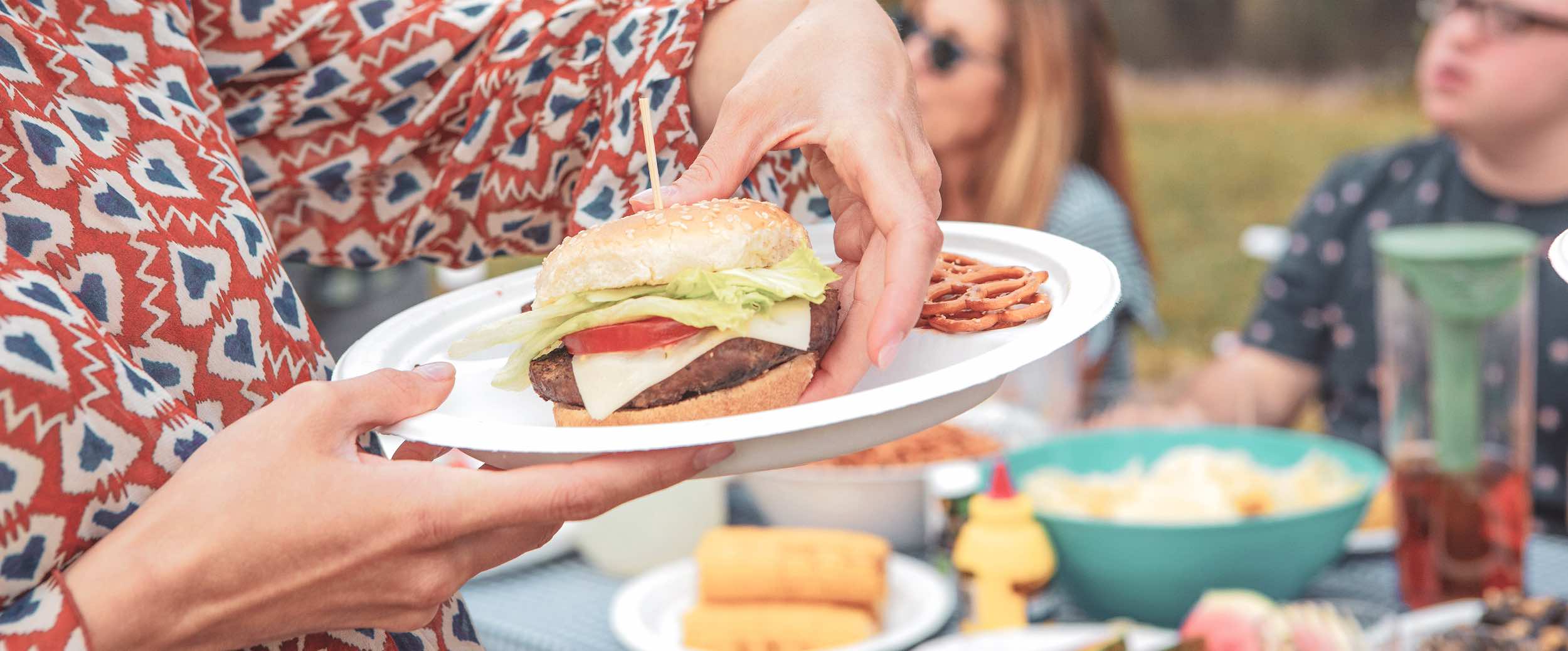 A woman at an outdoor barbecue picking a hamburger up off of a Chinet Classic plate