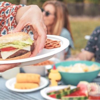 A woman at an outdoor barbecue picking a cheeseburger up off a Chinet Classic plate