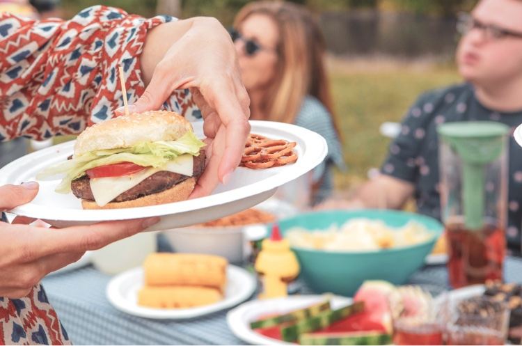 A woman at an outdoor barbecue picking a cheeseburger up off a Chinet Classic plate