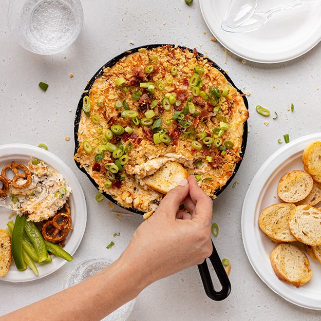 Hand scooping jalapeno popper dip from a cast iron pan with a tortilla chip