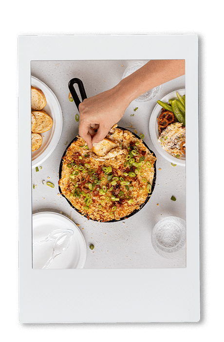 Instax picture of a hand scooping jalapeno popper dip from a cast iron pan with a tortilla chip