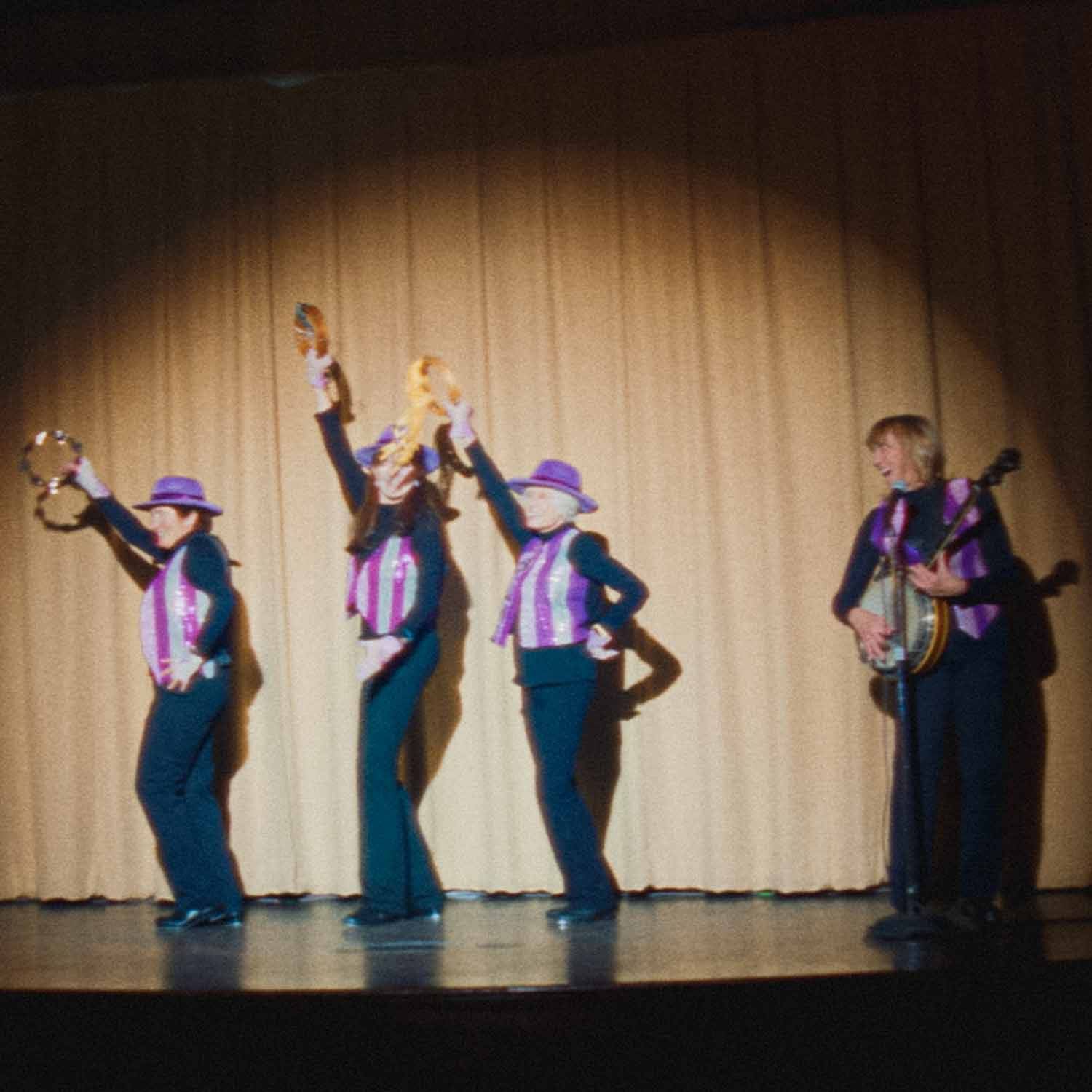 Tap Chicks performing with tambourines and a banjo in purple vests and hats on stage