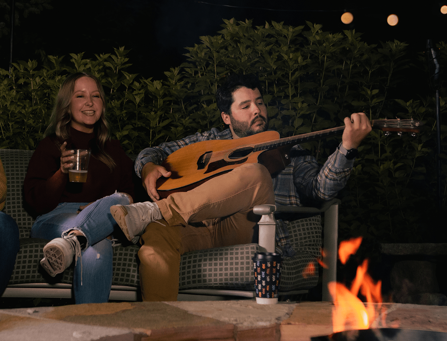 Man playing a guitar sitting next to a woman infront bonfire