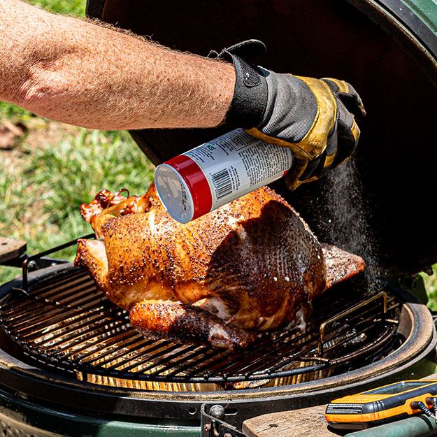 A hand spraying cooking spray on a turkey in a smoker