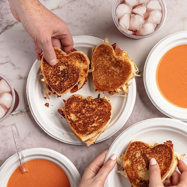 Hand grabbing a heart-shaped chicken bacon grilled cheese served on Chinet Classic plates served next to tomato soup served in Chinet Classic bowls