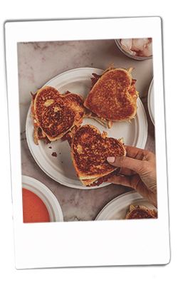 Instax picture of a hand grabbing a heart-shaped chicken bacon grilled cheese served on Chinet Classic plates served next to tomato soup served in Chinet Classic bowls