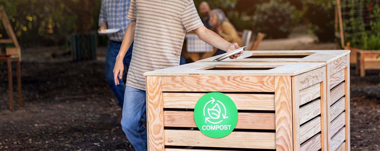 A person places a disposable plate into a wooden compost bin outdoors, marked with a green compost symbol, while other people stand nearby holding plates in a garden setting.