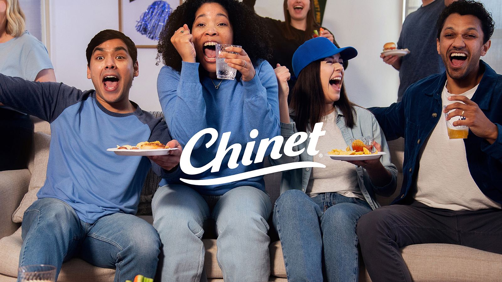 A group of friends sitting on a couch cheer excitedly while watching a game on TV, holding snacks and drinks on disposable plates; the white Chinet logo is prominently displayed in the center of the image.