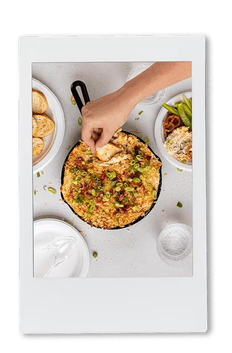 Instax picture of a hand scooping jalapeno popper dip from a cast iron pan with a tortilla chip