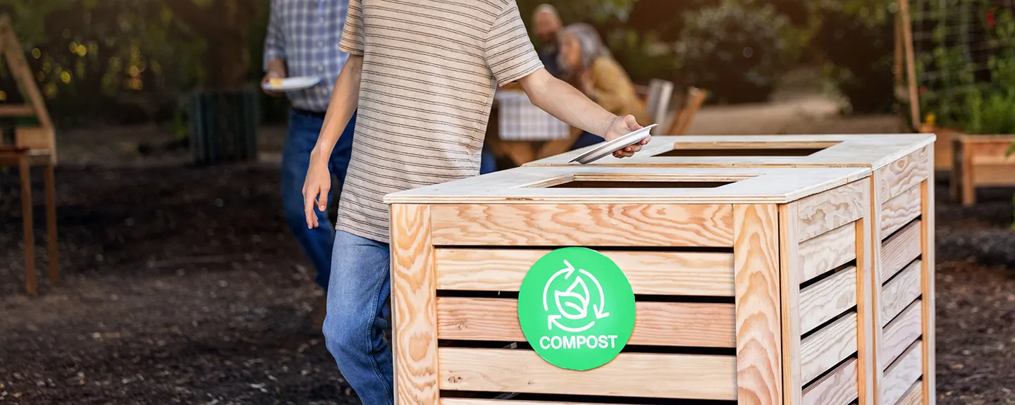 A person places a disposable plate into a wooden compost bin outdoors, marked with a green compost symbol, while other people stand nearby holding plates in a garden setting.